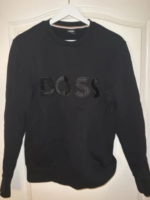 Svart sweatshirt från BOSS, storlek S - Svart sweatshirt från BOSS i storlek S med stort broderat BOSS-tryck på bröstet. Tröjan har rund hals, långa ärmar och en klassisk, normal passform. Materialet är mjukt och känns som bomull, perfekt för chill dagar.