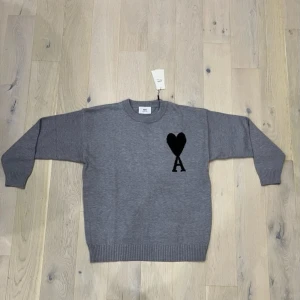 Grå stickad tröja från Ami - Snygg grå stickad tröja från Ami Alexandre Mattiussi med svart hjärta och bokstaven A på bröstet. Tröjan har rund hals, ribbade muddar och lång ärm. Perfekt för dig som gillar stilrena detaljer och vill ha något som sticker ut lite.