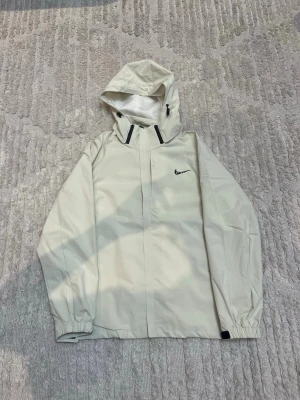 Beige regnjacka från Nike med huva - Säljer en beige regnjacka från Nike med huva och svarta detaljer vid dragkedjan. Jackan har elastiska muddar vid ärmslut och en stilren design med Nike-logga på bröstet. Perfekt för blöta dagar och enkel att matcha med din streetwear.