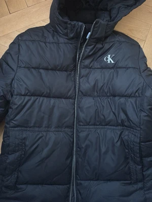 Svart pufferjacka från Calvin Klein Jeans - Svart pufferjacka från Calvin Klein Jeans med CK-logga på bröstet och ärmen. Jackan har huva, dragkedja framtill och två sidofickor. Perfekt för kalla dagar och har en clean, stilren look som funkar till jeans och sneakers.