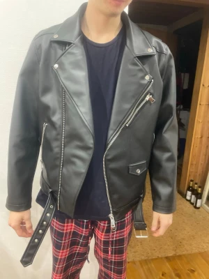 Svart skinnjacka från Jack & Jones - Säljer en svart skinnjacka från Jack & Jones i storlek L. Klassisk bikerstil med sned dragkedja, flera fickor med dragkedjor och ett bälte i midjan. Jackan har breda slag och silverfärgade detaljer som ger en cool look.