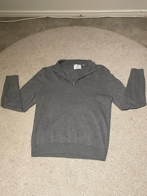 Grå half zip tröja från Bläck - Säljer en stilren grå tröja från Bläck med half zip-dragkedja och hög krage. Tröjan är långärmad och har en klassisk, enkel design som passar perfekt till lager på lager. Materialet känns mjukt och är troligtvis bomull eller en bomullsblandning.