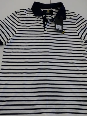 Randig polo från Lyle & Scott - Snygg randig polo från Lyle & Scott i offwhite och marinblått. Klassisk krage, korta ärmar och broderad gul logga på bröstet. Skön bomullskvalitet och stilrena knappar framtill. Perfekt för dig som gillar enkel men cool stil.