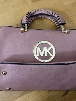 Rosa handväska från Michael Kors - Säljer en snygg rosa handväska från Michael Kors med stort MK-emblem i guld på framsidan. Väskan har ett handtag med dekorativa detaljer och guldfärgade metalldelar. Perfekt för dig som gillar stilrena accessoarer med lite lyxig känsla. Äkta men en väldigt gammal modell