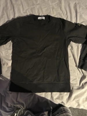Svart sweatshirt från Stone Island - Säljer en svart sweatshirt från Stone Island med den klassiska kompasspatchen på ärmen. Tröjan har rund halsringning, långa ärmar och ribbade muddar. Tillverkad i mjuk bomull för skön vardagsstil.