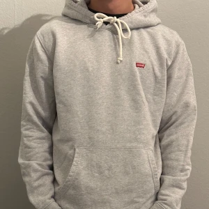 Grå hoodie från Levi's - Säljer en stilren grå hoodie från Levi's med en liten röd logga på bröstet. Tröjan har en klassisk känguruficka och justerbar huva med snören. Perfekt för en avslappnad look.