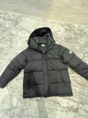 Burberry  - Svart pufferjacka från Burberry med huva och dragkedja samt knappar framtill. Jackan har två sidofickor med dragkedja och en patch med Burberry London England på ärmen. Insidan av huvan har ett grått och randigt foder. Perfekt för kalla dagar. 