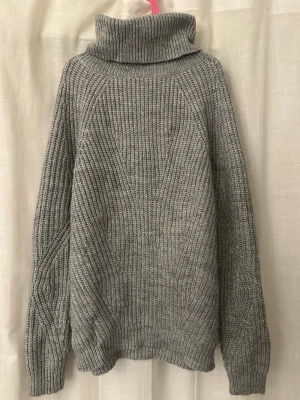 Grå stickad polotröja - Säljer en grå stickad polotröja med bred krage och långärmad design. Tröjan har ett grovt ribbat mönster och är perfekt för kyliga dagar. Passformen är normal och materialet känns mjukt och värmande.