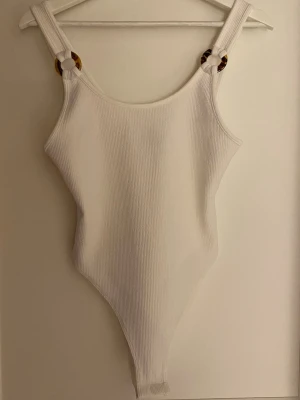 Vit ribbad linnetopp med ringdetaljer - Säljer en stilren vit ribbad linnetopp med breda axelband och snygga ringdetaljer i sköldpaddsmönster. Toppen har en kroppsnära passform och är perfekt till sommaren. Materialet är mjukt och stretchigt för en bekväm känsla.