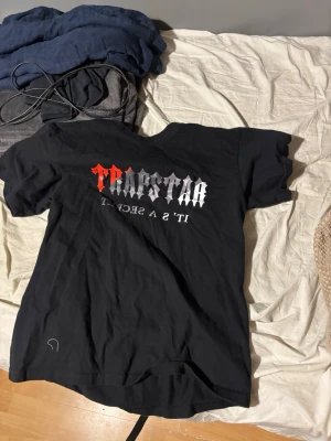 Svart Trapstar t-shirt med tryck - Svart t-shirt från Trapstar med stort tryck i rött och vitt på bröstet. Texten har en edgy, gotisk stil och t-shirten har en klassisk rund hals och korta ärmar. Materialet känns mjukt och är troligtvis bomull, perfekt för en streetwear-look.