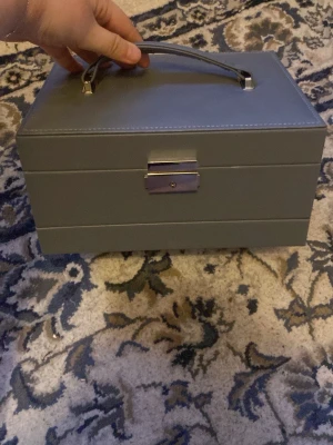 Grå smyckesväska med spegel - Snygg och stilren grå smyckesväska i syntetmaterial med handtag och metallspänne. Väskan har flera fack och en inbyggd spegel, perfekt för att organisera ringar, halsband och örhängen. Enkel att bära med sig och passar bra på sminkbordet. Innehåller följer INTE med