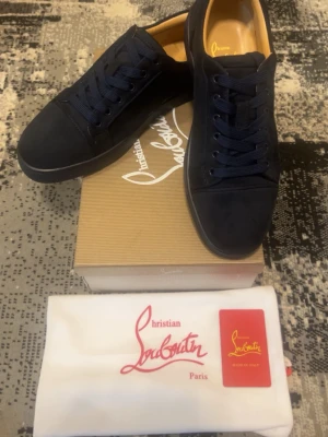 Mörkblå sneakers från Christian Louboutin - Snygga mörkblå sneakers från Christian Louboutin med klassisk röd sula och rund tå. Skorna har snörning och är tillverkade i mocka med detaljer i skinn. Kommer med originalkartong och dustbag. Perfekt för dig som gillar exklusiva märken och stilren design.