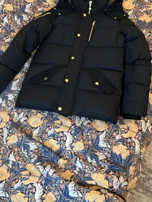 Svart pufferjacka Moose Knuckles M - Säljer min moose knuckles jacka, som är köpt för 11000, vilket jag har kvitto på som jag nu säkjer för 7000 Svart pufferjacka från Moose Knuckles med guldiga detaljer och dragkedjor. Jackan har huva, flera fickor med lock och snygga knappar. Materialet är polyester och dun, perfekt för kalla dagar. Passformen är normal och sitter perfekt på storlek S.