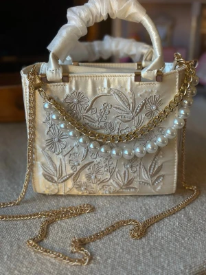 Broderad vit handväska med pärlor - Snygg vit handväska i satin med blommigt broderi och pärldetaljer. Väskan har guldfärgad kedja som axelrem och pärlor framtill. Handtaget är inslaget i tyg och väskan har en boxig form. Perfekt för dig som gillar detaljer och vill sticka ut. NO BOX BAG ONLY