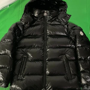 Svart Moncler Maya liknande pufferjacka - Alla storlekar finns, kom priv. Leverans tar ca 2 veckor!  Snygg svart pufferjacka från Moncler med glansig finish och huva. Jackan har två fickor med dragkedja framtill och tydliga Moncler-detaljer på ärmen och dragkedjan. Perfekt för kalla dagar när du vill hålla stilen.