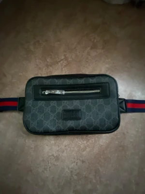 Gucci svart magväska med monogram - Snygg svart magväska från Gucci med klassiskt GG-monogram över hela väskan. Väskan har en silverfärgad dragkedja framtill och ett justerbart band i marinblått och rött. Perfekt för att bära det viktigaste på ett stilrent sätt.