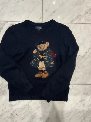 Mörkblå stickad tröja Polo Bear - Säljer en mörkblå stickad tröja från Polo Ralph Lauren med ikonisk Polo Bear-motiv framtill. Tröjan har rund hals, långa ärmar och RL-broderi i rött vid nederkanten. Perfekt för dig som gillar klassisk stil med en twist.