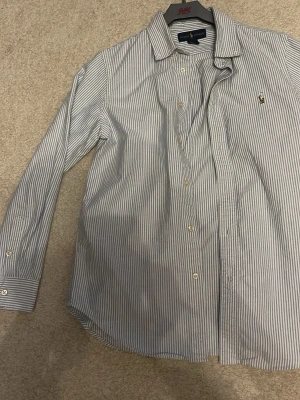 Randig skjorta från Ralph Lauren - Klassisk skjorta från Ralph Lauren i vit och blå smalrandig design. Skjortan har lång ärm, knappar framtill och den ikoniska broderade loggan på bröstet. Tillverkad i mjuk bomull och har en snygg krage som ger en clean look.