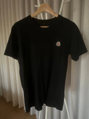 Svart t-shirt från Moncler - Svart t-shirt från Moncler med klassisk rund hals och korta ärmar. Diskret Moncler-logga på bröstet ger en clean och stilren vibe. Tillverkad i mjuk bomull som känns skön mot huden. Perfekt för dig som gillar minimalistisk design och premiumkänsla.