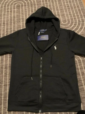 Svart hoodie från Polo Ralph Lauren - Svart hoodie med dragkedja från Polo Ralph Lauren. Klassisk design med huva och dragsko, två fickor fram och broderad logga på bröstet. Tillverkad i mjukt bomullsmaterial som är skönt mot huden. Perfekt för chill dagar och streetwear-stil.