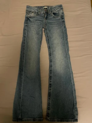 Blå bootcut jeans från Gina Tricot - Snygga blå jeans från Gina Tricot i bootcut-modell med klassisk femficksdesign och dubbla silverfärgade knappar fram. Jeansen har ljus tvätt och kontrastsömmar, tillverkade i jeansmaterial som ger en cool och avslappnad vibe.