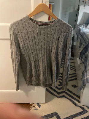 Grå kabelstickad tröja Polo Ralph Lauren - Snygg grå kabelstickad tröja från Polo Ralph Lauren i barnstorlek 10-12 år. Tröjan har rund hals, långärmade och ett broderat svart Polo-logo på bröstet. Ribbstickade muddar vid ärmslut och nederkant. Perfekt för kyliga dagar.