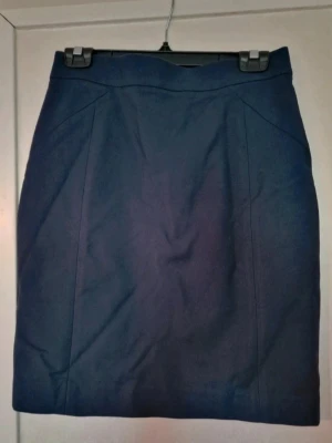 Kort marinblå kjol med slits - Snygg mörkblå kjol med klassisk, rak form och diskret slits bak. Kjolen har en dold dragkedja i ryggen. Kjolen är i mycket bra skick. Den skavank jag har kunnat hitta är någon lös tråd vid slitsen (se bild). 