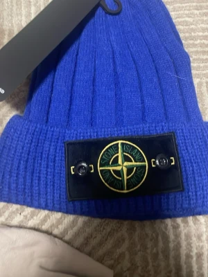 Blå stickad mössa från Stone Island - Säljer en snygg blå stickad mössa från Stone Island med klassisk logotyp framtill. Mössan har bred uppvikt kant och ribbad struktur som ger en schysst look. Perfekt för dig som vill ha streetstyle och värme i vinter.