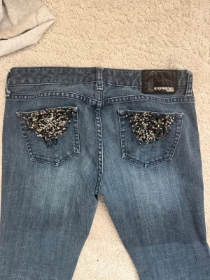Express Stella jeans med paljetter - Snygga blå jeans från Express, modell Stella, med låg midja och bootcut. Baktill har fickorna coola paljett-detaljer i svart och silver som ger extra bling. Jeansen är gjorda i klassiskt denim och har en skön passform som sitter bra över höften. Midjemått: ca 39,5cm och inerbenslängd: ca 77cm