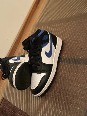 Nike Air Jordan 1 svart/blå sneakers äkta  - Nike Air Jordan 1 sneakers i svart, vit och blå med klassisk hög siluett. Skorna har svart snörning, rund tå och platt sula. Swoosh-loggan i blått på sidan och Air Jordan-märket på ankeln. Tillverkade i skinn med perforerad tå för extra ventilation. Andvänd 2 gånger! 