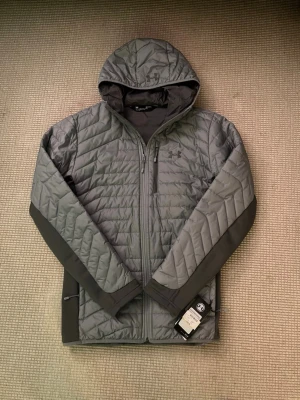 Grå quiltad pufferjacka Under Armour - Quiltad pufferjacka från Under Armour i grått med huva och hel dragkedja. Jackan har snygga paneler längs ärmarna och diskret logga på bröstet. Fitted passform och tillverkad i syntetmaterial, perfekt för kyliga dagar. Originella pris 1399kr som ni ser på bilden kommer sälja den för 845kr