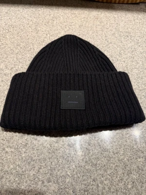 Svart mössa från Acne Studios i ull - Ribbstickad svart mössa från Acne Studios med ikonisk ansiktslogga framtill. Tillverkad i 100% ull för extra värme och stil. Klassisk vikt kant och diskret Acne Studios-tag på insidan.