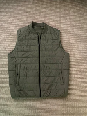 Olivgrön quiltad dunväst från Zara - Säljer en olivgrön quiltad dunväst från Zara i storlek M. Västen har en rak passform, ståkrage och praktisk dragkedja framtill. Två sidofickor med diskreta knappar och horisontella sömmar ger en clean look. Perfekt att slänga över en hoodie eller tröja.