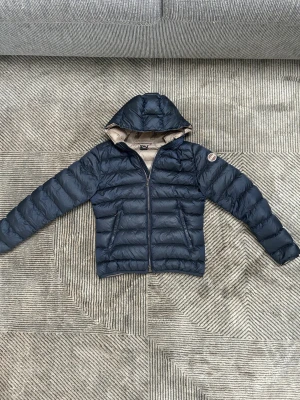 Mörkblå Colmar dunjacka  - Snygg mörkblå dunjacka från Colmar med beige insida och huva. Jackan har ett quiltat mönster, dragkedja framtill och två sidofickor. Märkespatch på ärmen och diskret logga bak. Perfekt för kalla dagar och riktigt stilren look.