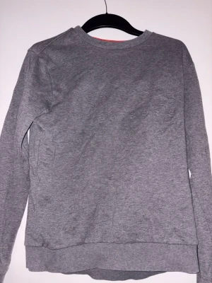 Grå långärmad tröja från Hugo Boss - Snygg grå långärmad tröja från Hugo Boss med diskret broderad logga på bröstet. Tröjan har rund hals och ribbade muddar vid ärmslut och nederkant. Passformen är slim fit och insidan av kragen har en cool röd detalj. Perfekt för dig som gillar stilrena plagg.