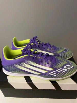 Adidas F50 Club In lila fotbollsskor (45,5) - Adidas F50 Club fotbollsskor i en snygg lila färg med neon-gula detaljer och vita ränder. Skorna har snörning och en platt sula, designade för inomhusspel. Ovandelen är tillverkad i syntetmaterial med ett grafiskt mönster och texten F50 på sidan.
