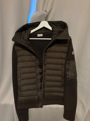 Moncler cardigan - Säljer en grön cardigan från Moncler med stickade ärmar och huva. Jackan har quiltad front, dragkedja och en ficka med zip på ärmen. Perfekt för dig som vill ha en snygg och chill look med exklusiv känsla. Storlek M men kan passa S. 