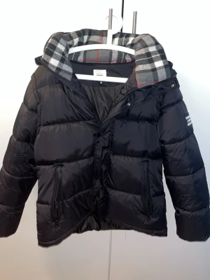 Svart pufferjacka från Burberry - Svart pufferjacka från Burberry med huva och klassiskt rutigt foder. Jackan har en stor Burberry London England-patch på ärmen, dragkedjor på fickorna och quiltad design. Perfekt för kalla dagar och har en riktigt snygg streetstil. XS men sitter som S.