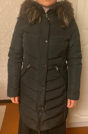 Svart lång dunjacka med päls - Säljer en svart lång dunjacka med fluffig päls runt huvan. Jackan har quiltad design, dragkedja och knappar framtill samt två snedställda fickor med dragkedja. Perfekt för kalla dagar och har en snygg, figurnära passform.