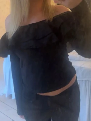 Svart offshoulder volangtop - Säljer en svart offshoulder topp med volangdetalj över bröstet. Toppen har lång ärm och är croppad så magen syns. Perfekt för dig som gillar trendiga och stilrena plagg med lite extra edge.