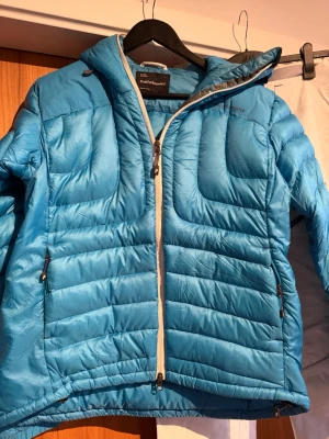 Blå dunjacka Peak Performance dam L - Säljer en blå dunjacka från Peak Performance i dammodell, storlek L. Jackan har quiltad design, huva och vita dragkedjor framtill och på fickorna. Perfekt för kalla dagar och riktigt snygg färg som sticker ut i vintermörkret.
