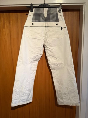 Vita täckbyxor Peak Performance dam - Säljer ett par vita täckbyxor från Peak Performance för dam. Byxorna har flera dragkedjeförsedda fickor, bälteshällor och coola detaljer i svart och grått upptill. Perfekta för vinterns alla äventyr i backen eller snön.