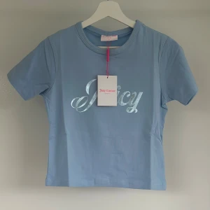 Blå Juicy Couture t-shirt i bomull - Jättefin ljusblå sport T-shirt från Juicy Couture med glansigt tryck framtill där det står 'Juicy Couture' i metallicblått och silver. Ny med etikett!. Se bild för mått.  
