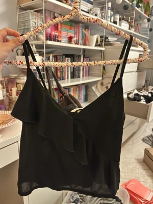 Svart linnetopp med volang - Säljer en svart linnetopp med smala axelband och snygg volangdetalj framtill. Toppen är ärmlös och har en lätt, flowy passform som känns luftig och skön. Perfekt att styla med jeans eller kjol för en cool look.