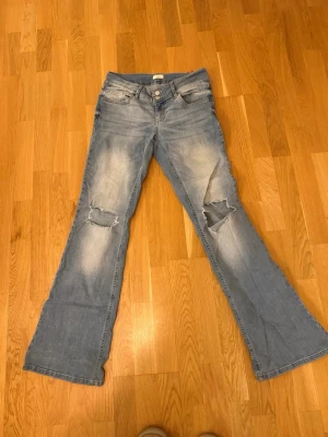 Bootcut ljusblå jeans med slitningar - Säljer ett par ljusblå bootcut jeans med slitna detaljer på knäna, en bältes hållare är lös och de är väl använda.  köpta på nelly 