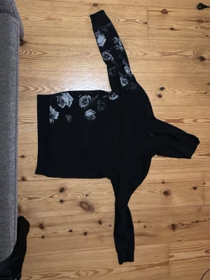 Svart/Vit hoodie från H&M med rosor på. - Svart Hoodie från H&M, vita roser som rör sig upp från den stora uppningen till höger armen.