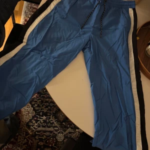 Blå trackpants från Burberry - Säljer ett par blå täckbyxor från Burberry med svarta och vita paneler längs sidorna. Byxorna har snörning i midjan och tryckknappar på sidan. Materialet är syntet och de har en sportig vibe med Burberry-logga broderad vid benet.