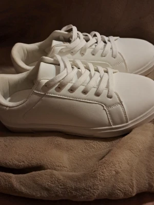 Vita sneakers med snörning - Fräscha vita sneakers med klassisk rund tå och platt sula. Skorna har vit snörning och är tillverkade i syntetmaterial med mjukt foder. Perfekt för dig som gillar stilrena och enkla sneakers som funkar till många outfits.