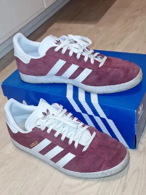 Adidas Gazelle vinröda sneakers - Säljer ett par Adidas Gazelle sneakers i vinrött med klassiska vita ränder och vit sula. Skorna har snörning och är tillverkade i mocka med detaljer i syntet. Ikonisk retrostil som passar perfekt till streetwear och vardagsoutfits. Levereras i originalkartong. Använda endast fåtal gånger.