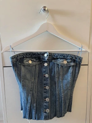 Blå denim korsett-topp från Zara - Säljer en blå denim korsett-topp med silverfärgade knappar framtill och två dekorativa fickor. Toppen är axelbandslös och har en smockad baksida för extra komfort. Köpt på Zara och säljer då den inte kommer till använding💕💕